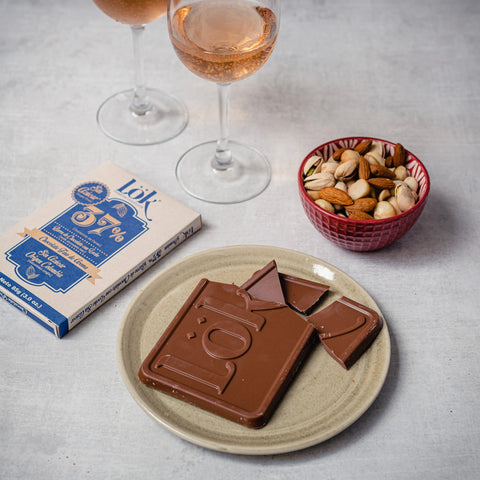 A styled product shot of a bar of Lok 52% dark chocolate on a beige plate with broken pieces, alongside a box of Lok chocolate, a bowl of mixed nuts, and two glasses of rosé wine on a light gray surface.