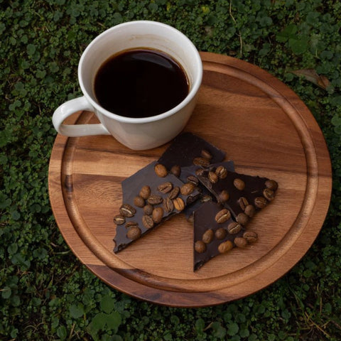 White mug of coffee on a wooden tray with chocolate and coffee beans, set against a green grass background.